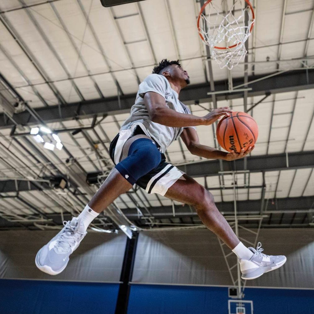 A basketball player in mid-air performing a dunk, wearing a blue knee brace on his right leg.