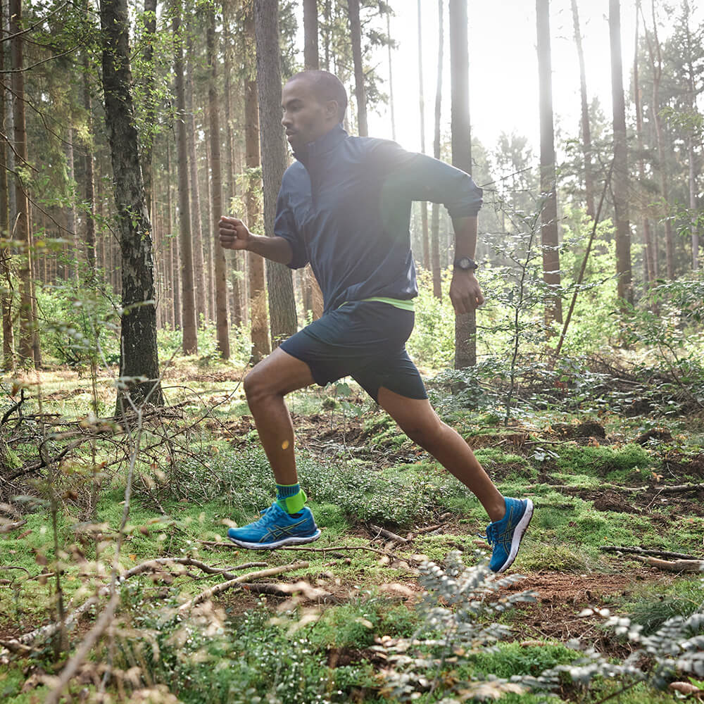 A man runs dynamically through a sunny forest. He wears a blue jacket, black shorts, and bright green calf sleeves.