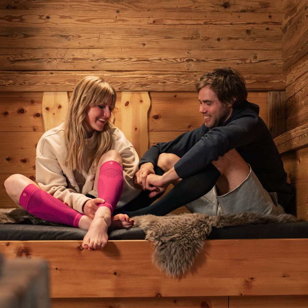 A smiling couple sits comfortably in a wooden cabin on a bench with a fur cover. The woman is wearing pink compression sleeves, while the man is wearing black ones. Both are enjoying their well-earned rest after an active day.