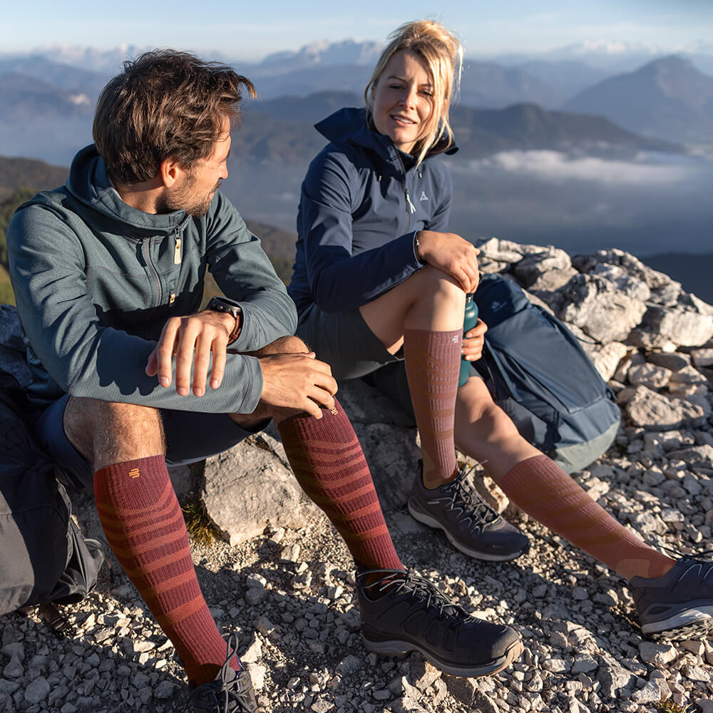 Two hikers enjoying the view on a mountain summit while wearing Merino Outdoor Compression Socks in red-brown. The socks provide optimal comfort and support for long treks in challenging terrain.