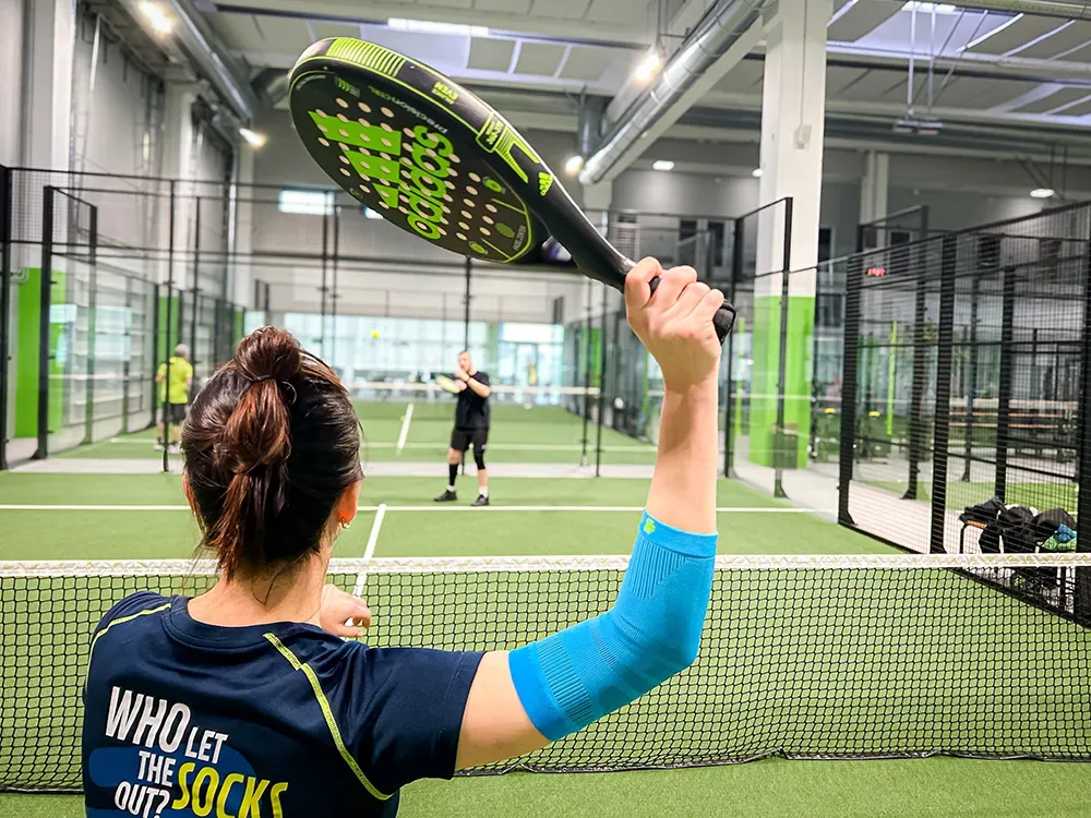 A padel tennis player stands at the net, preparing to hit the ball. Her arm is supported by a light blue compression sleeve.