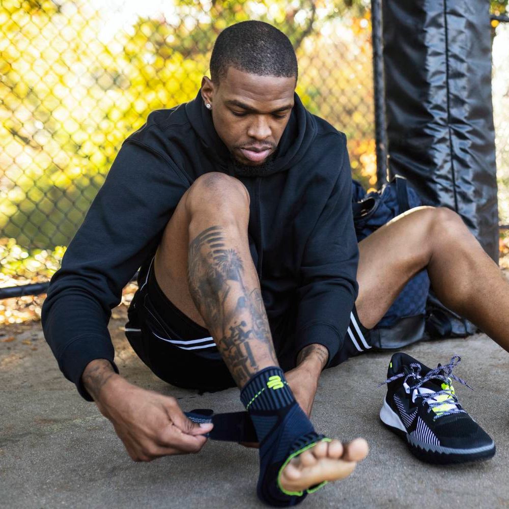 A sportsman sits outdoors, wrapping a Bauerfeind Sports Ankle Support brace around his ankle. His sports shoes are nearby, with a metal fence in the background.