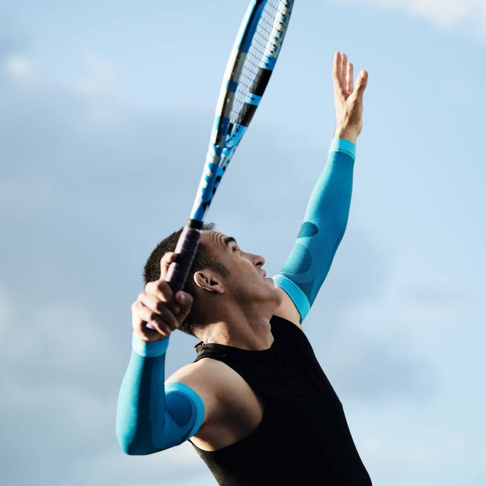 A tennis player wearing blue compression sleeves serving. The sky is blue with a few clouds.