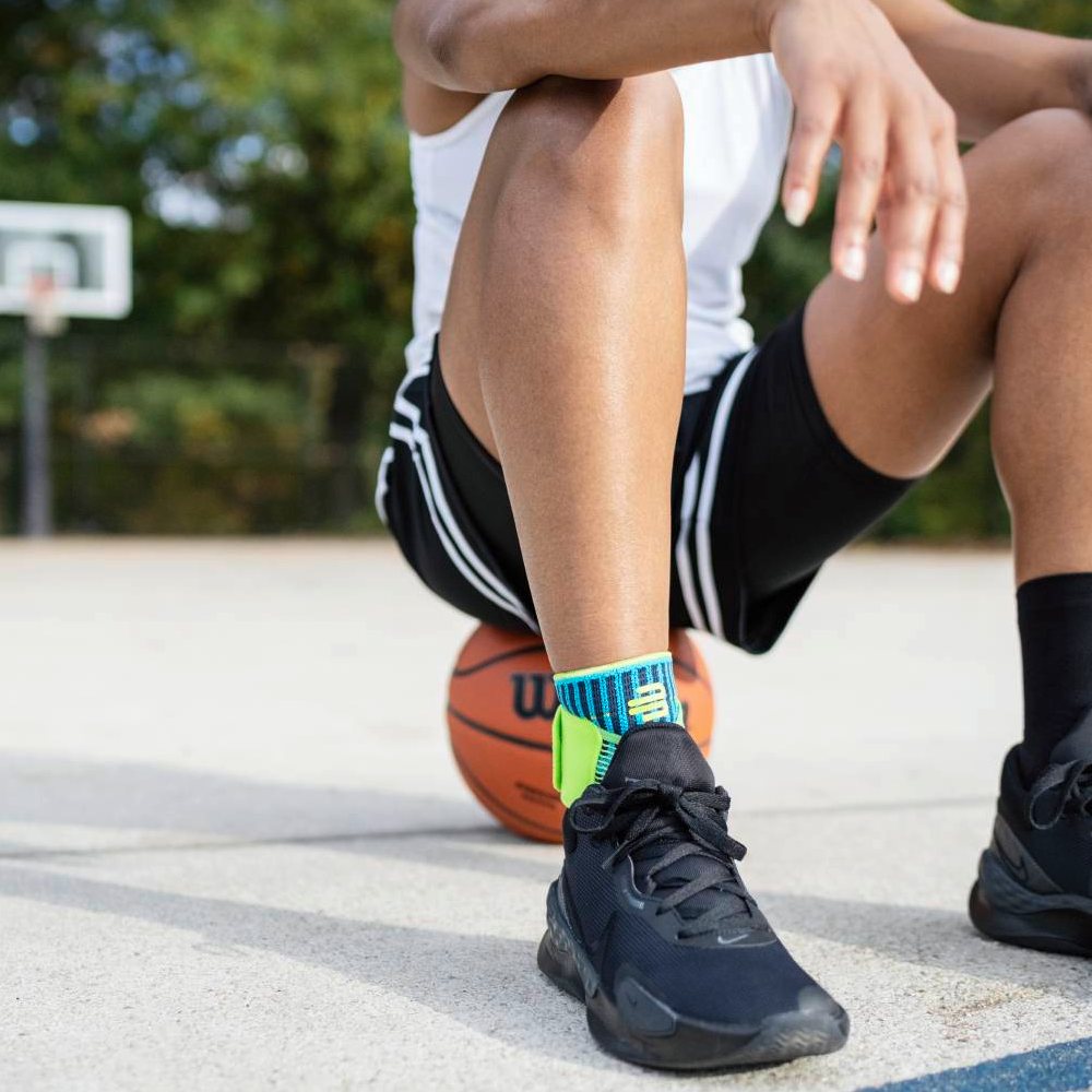 A basketball player sits on the court with a basketball under his leg. He is wearing the Bauerfeind Sports Ankle Support Rivera in blue-green on his ankle.