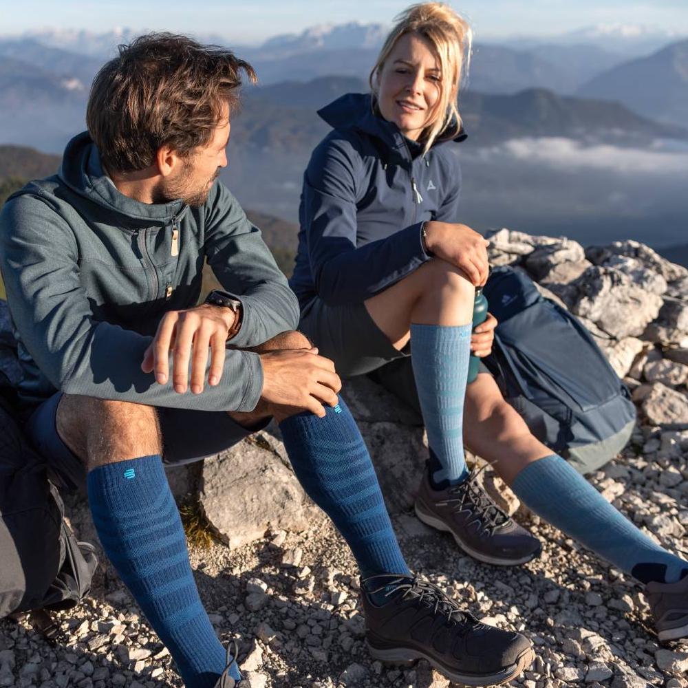 Two hikers enjoying the view on a mountain summit while wearing Merino Outdoor Compression Socks in red-brown. The socks provide optimal comfort and support for long treks in challenging terrain.