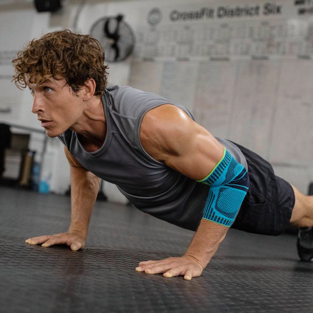 A fit man with a muscular upper body performs push-ups in a gym with focus. He is wearing a blue and black elbow support on his right arm for stability and support during his workout.