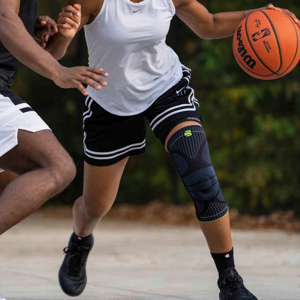 A female basketball player wears a black knee support on her right leg while moving dynamically with the ball past a defender.