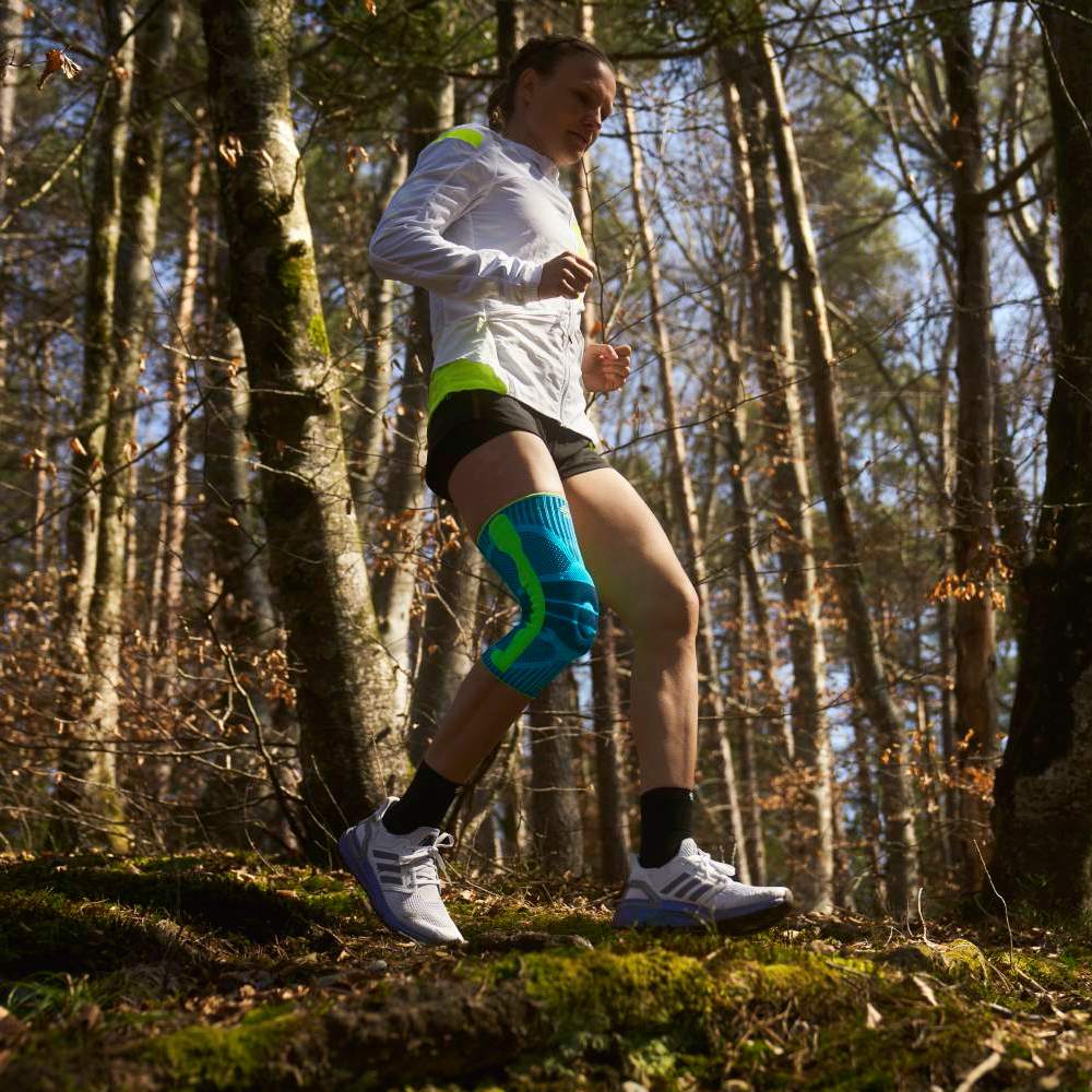 A woman runs along a forest trail in daylight, wearing a bright turquoise knee support on her left leg – focus is on movement and product use in an outdoor setting.