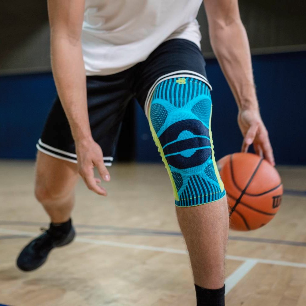 A basketball player wearing a turquoise knee support on his right leg dribbles the ball indoors, with the focus on the product and the athletic stance.