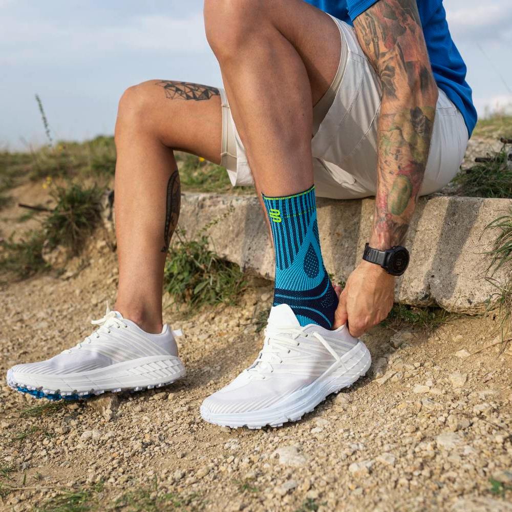 A man sits on a concrete ledge in a natural setting with a gravel path and grass. He wears white running shoes and blue sports socks with a striking pattern. His hands pull the shoe over the sock, while his tattooed arm is visible.