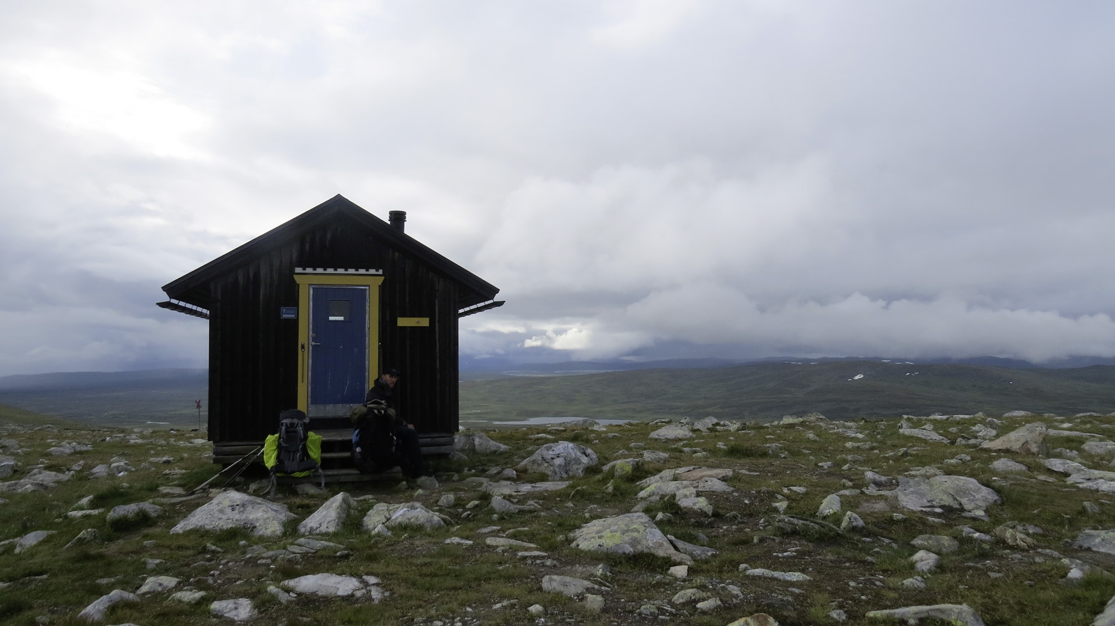 A lonely hut in the mountains in the middle of groving meadows with small rocks in the background the dense cloud cover and a small mountain lake