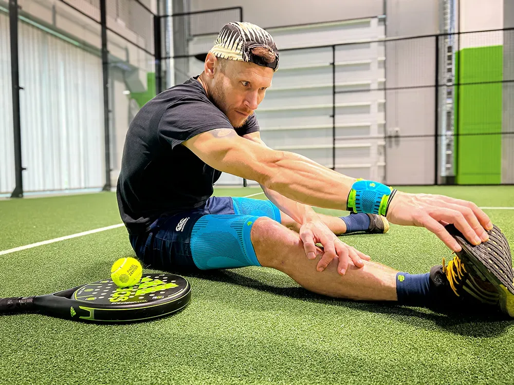 An athlete sits on the ground of a padel tennis court, stretching his leg muscles while wearing various compression products on his arm, leg, and wrist. A padel racket and a ball are placed beside him.