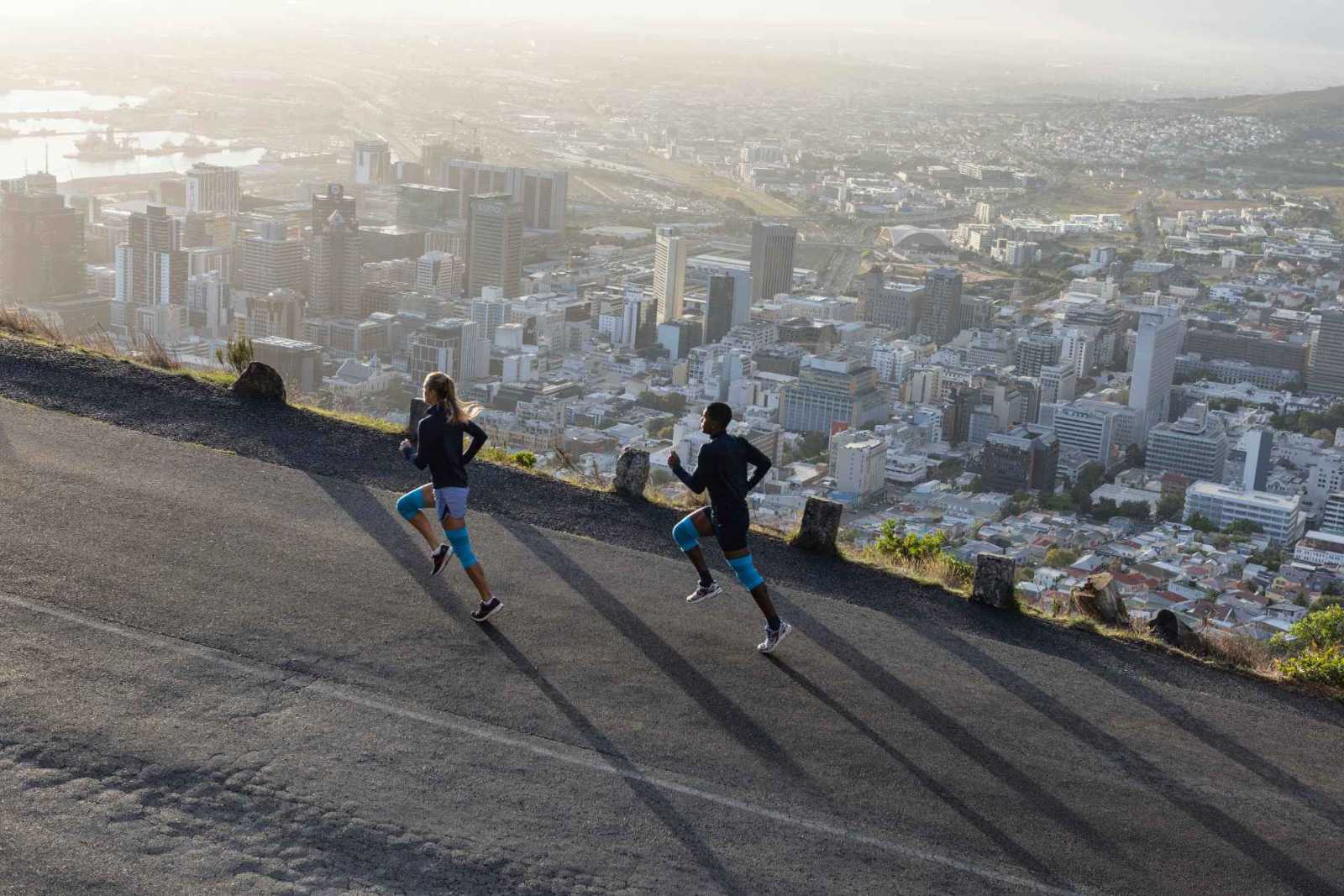 Very relaxed above the metropolis thanks to the Bauerfeind Sports Compression Knee Support A woman and a man with blue knee bars run up a street above a big city