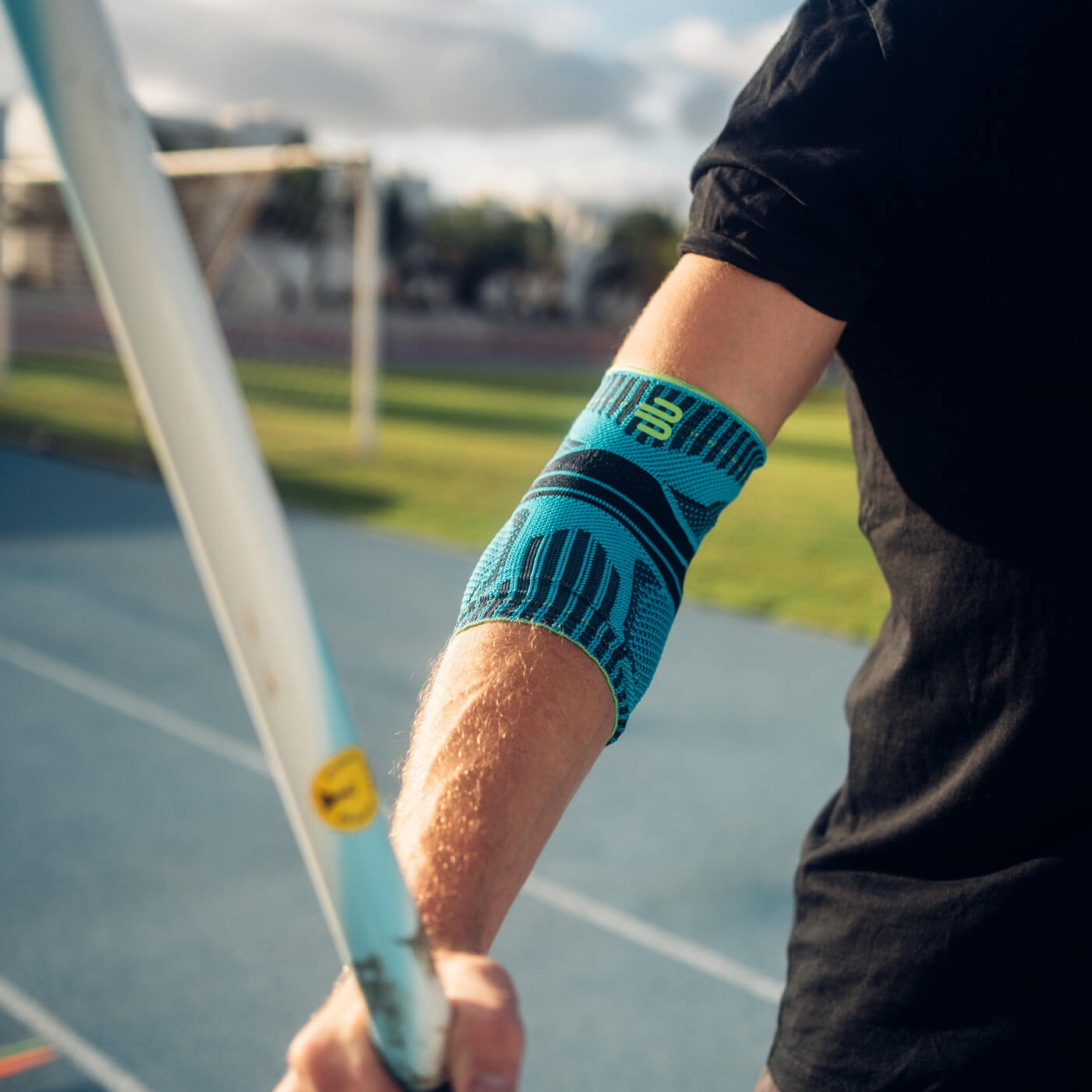 Close-up of an arm wearing a blue elbow support during a javelin throw. A running track and a sunny landscape are visible in the background.