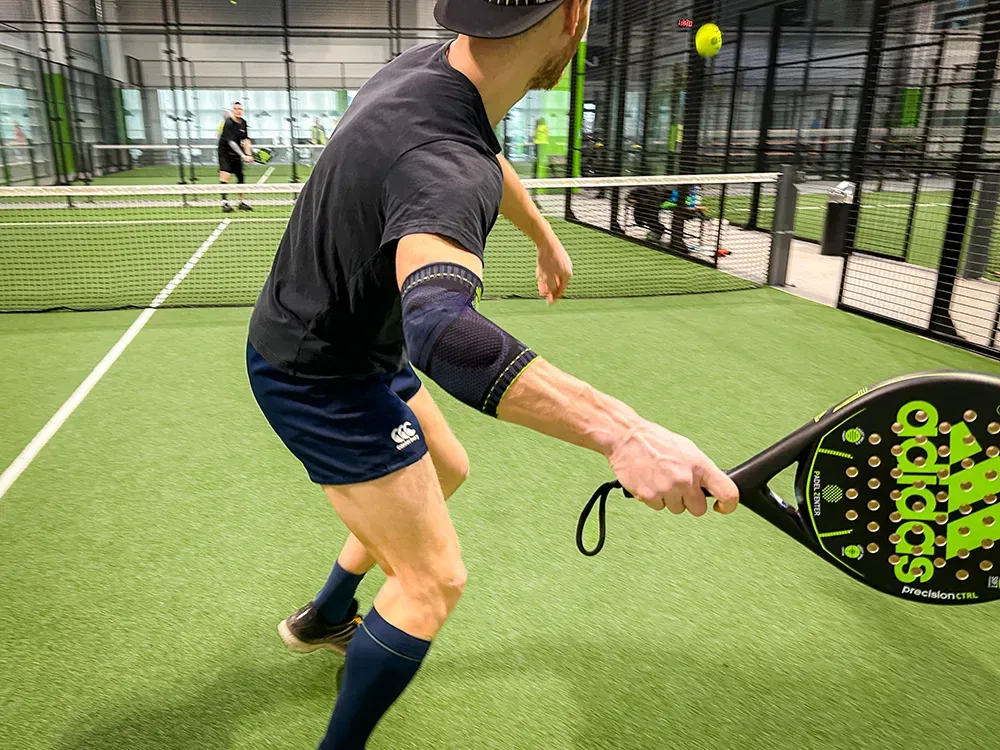 A padel tennis player in a black shirt and shorts hits the ball with full focus. He wears a dark blue sports support on his right arm for extra stability.