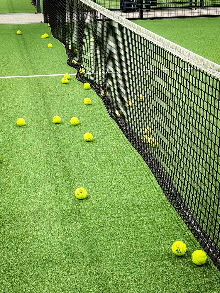 The court tells the story of a thrilling padel match. Several yellow padel balls are scattered across the green artificial turf court right next to the net – capturing the vibrant energy after intense rallies.