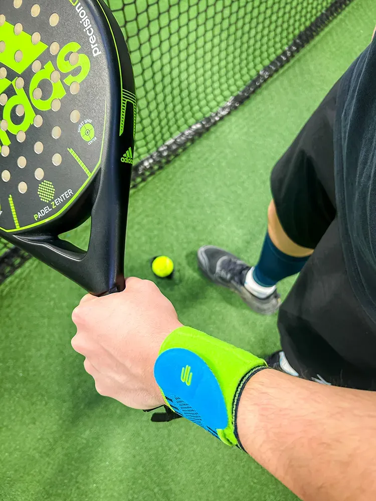 Close-up of a padel player holding an Adidas racket on the court. The athlete wears a neon wrist support, with a ball lying on the ground.