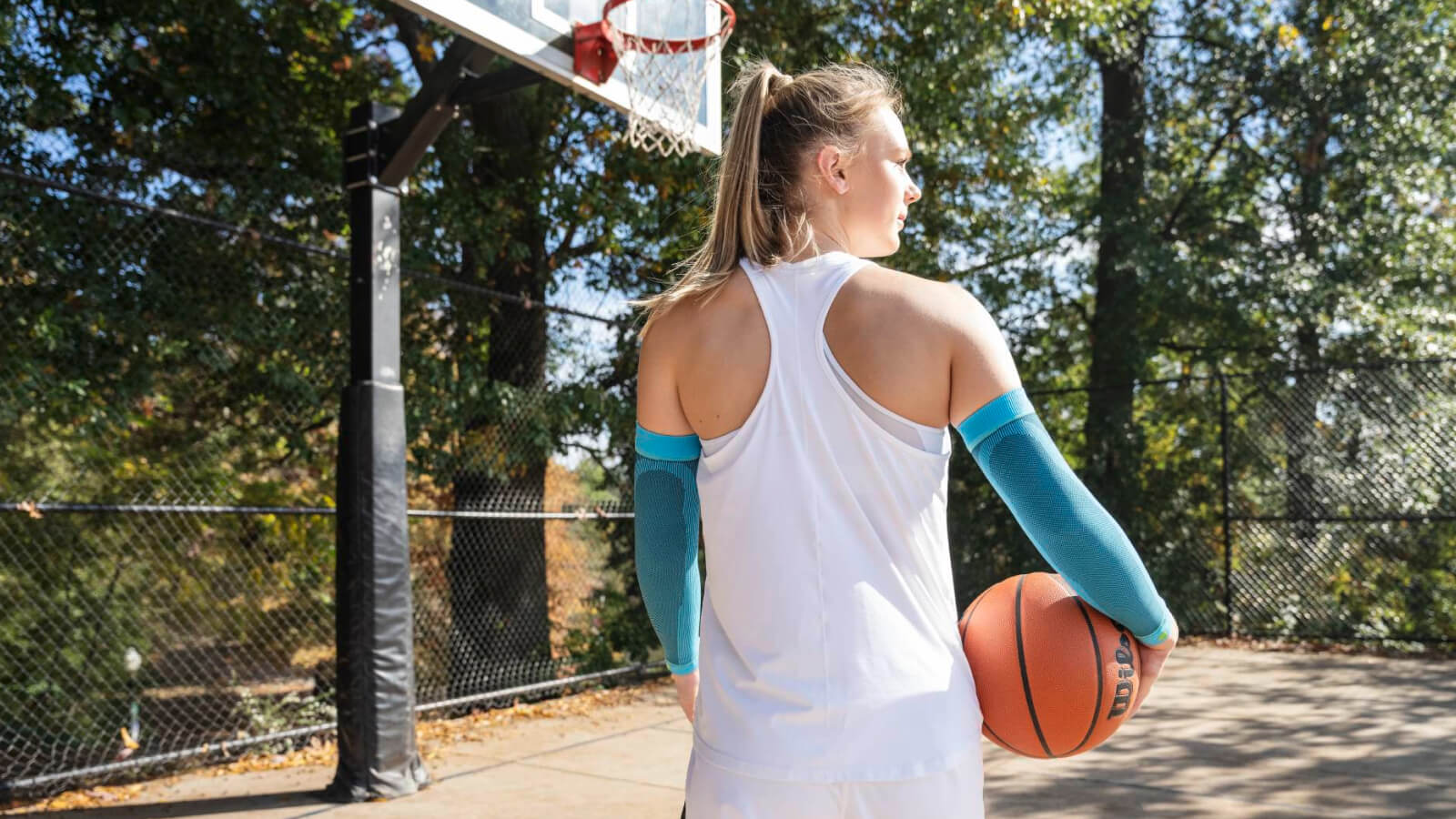 Ready for the next shot. A female basketball player stands on an outdoor court, holding a basketball in her right hand. She is wearing light blue compression arm sleeves and looking focused toward the hoop.