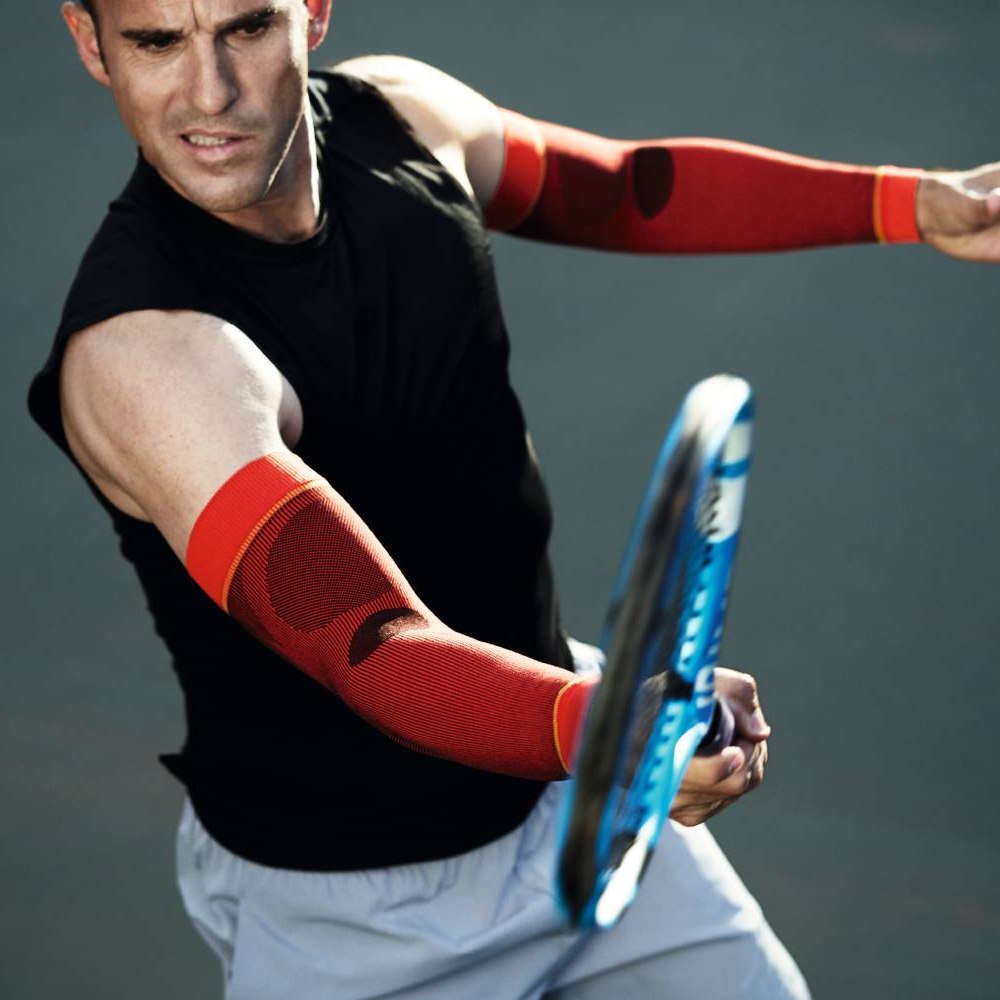 A tennis player wearing red compression sleeves hitting a ball with intense focus.