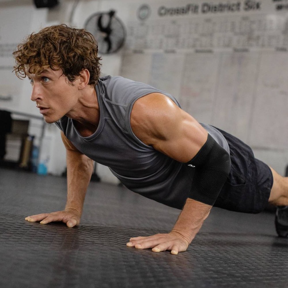 An athlete in the gym performing an intense push-up workout, wearing a black elbow support brace. The brace provides stable support and optimal protection for the elbow during strength training and athletic challenges.